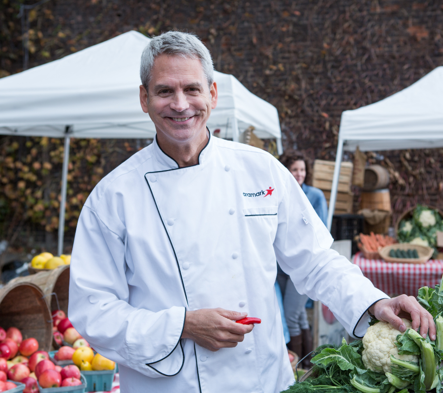 Aramark chef smiling with fresh vegetables at an outdoor farmers market.
